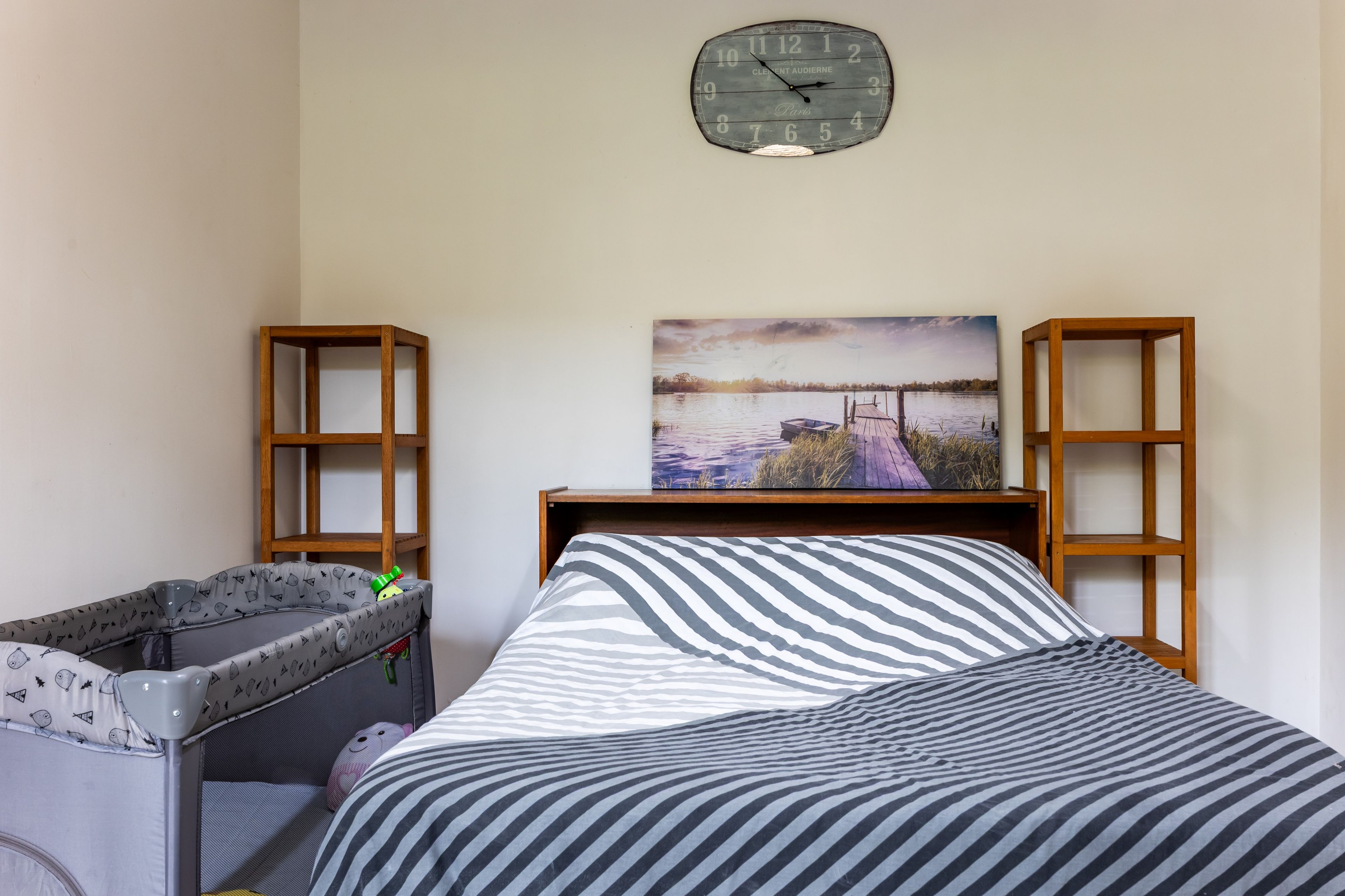 Une chambre à coucher avec un lit, un berceau, un tableau représentant un paysage au bord d’un lac et une horloge au mur.
