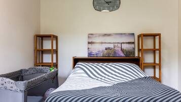 Une chambre à coucher avec un lit, un berceau, un tableau représentant un paysage au bord d’un lac et une horloge au mur.