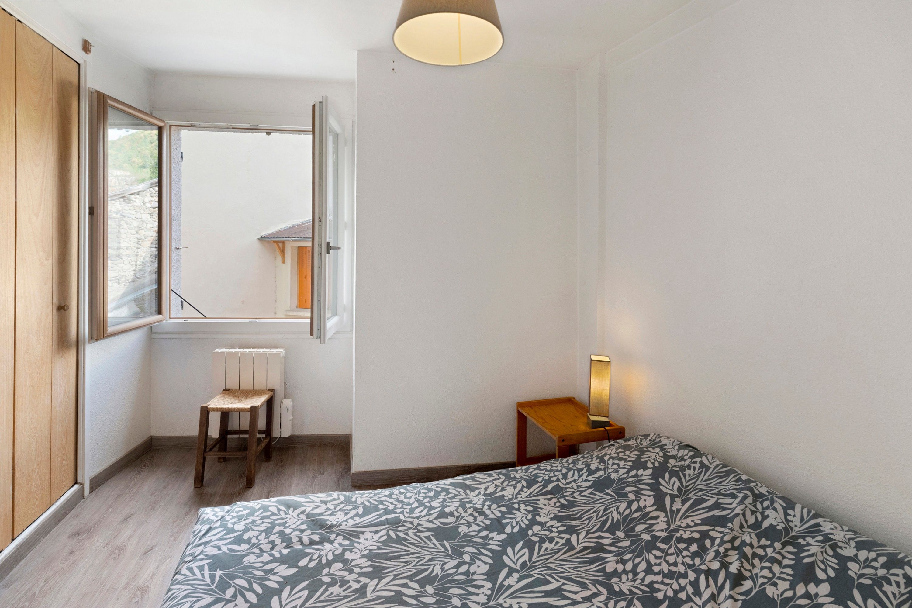Et soveværelse med seng, stol, natbord, lampe, vindue og radiator.