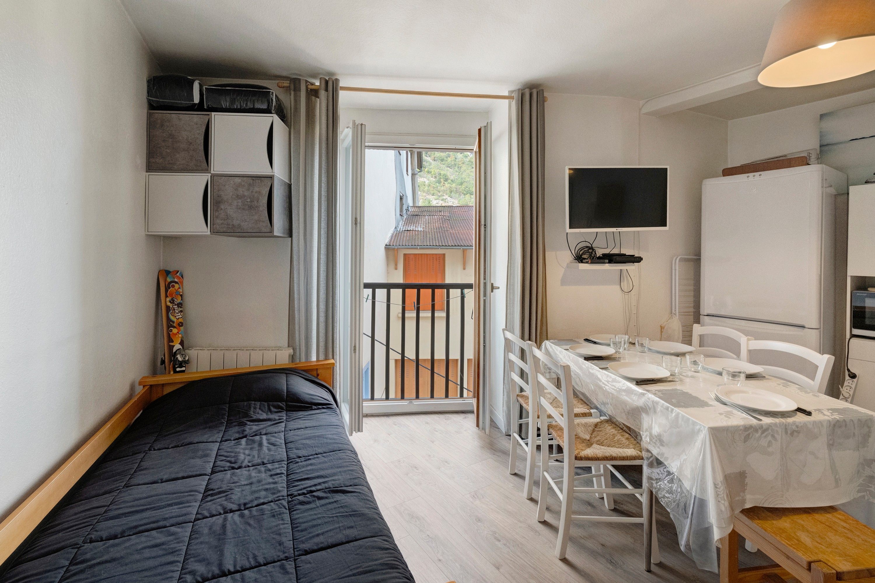Una habitación compacta con una cama, una mesa de comedor para dos personas, un televisor y un refrigerador.