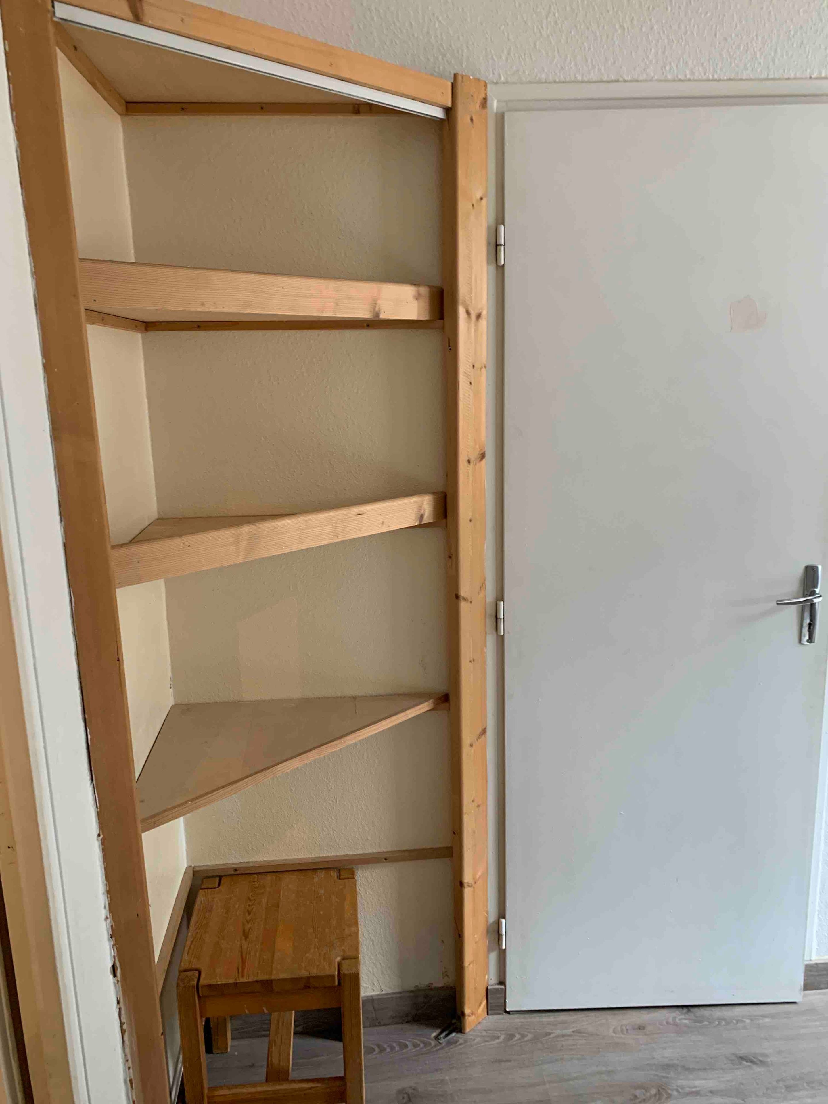 Una habitación con una estantería de madera contra la pared, un taburete pequeño de madera y una puerta blanca.
