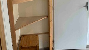 Una habitación con una estantería de madera contra la pared, un taburete pequeño de madera y una puerta blanca.