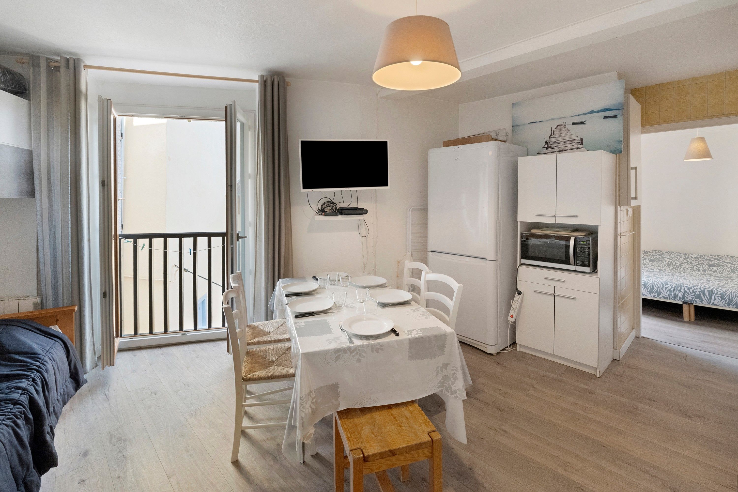 A compact living space with a dining area, a kitchenette, a TV mounted on the wall, and a balcony with a view.