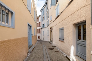 Un estrecho callejón entre edificios con un sendero de grava, puertas y ventanas con contraventanas.