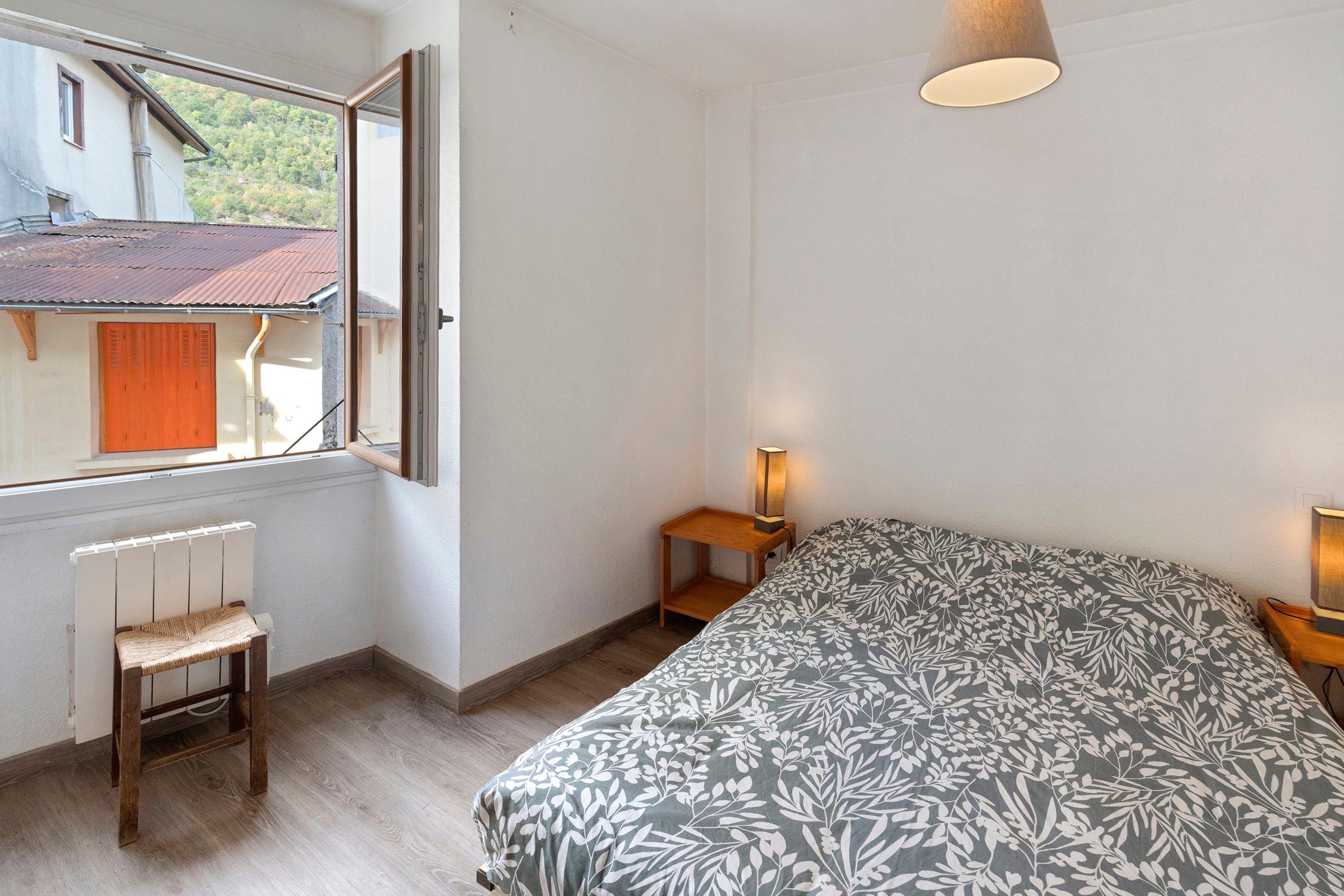 Un dormitorio con cama, una silla, una mesita de noche, una lámpara y una ventana con vista a una casa y árboles.