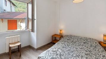 Un dormitorio con cama, una silla, una mesita de noche, una lámpara y una ventana con vista a una casa y árboles.