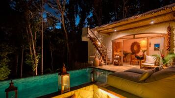 Un área junto a la piscina con un sofá, una escalera que sube a un nivel superior y un comedor con mesa y sillas.