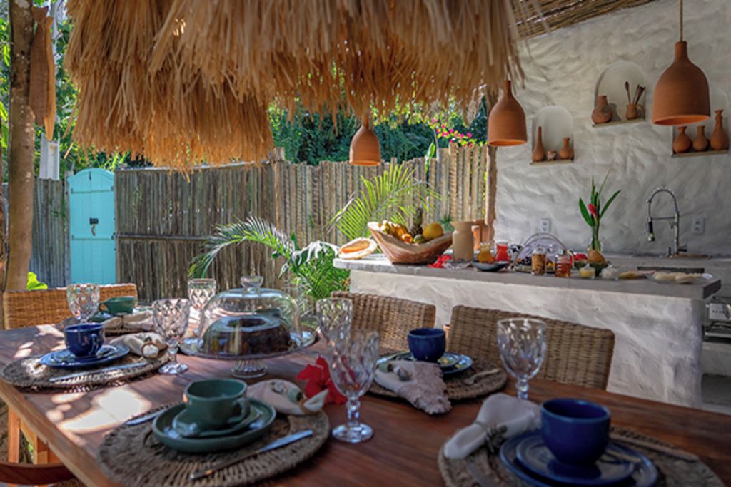 An outdoor dining area with a wooden table set for a meal, surrounded by wicker chairs, and a thatched roof overhead.