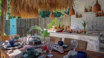 Uma área de refeições ao ar livre com mesa de madeira posta para uma refeição, rodeada por cadeiras de vime e um telhado de palha.