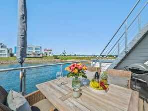 Une table ornée d’un vase de fleurs, d’une bouteille et d’un panier de fruits, placée sur le pont d’un bateau, avec vue sur les bâtiments et l’eau.
