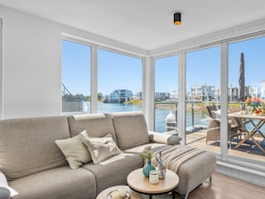 A modern living room with a large window overlooking a river, a sofa, a round coffee table, and a dining area with outdoor seating. - Holiday Home in Olpenitz With Swimming Pool (Kappeln)
