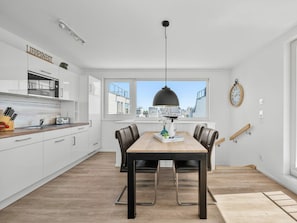 A modern kitchen with white cabinetry, a dining table with chairs, a pendant light, and a city view through large windows. - Holiday Home in Olpenitz With Swimming Pool (Kappeln)