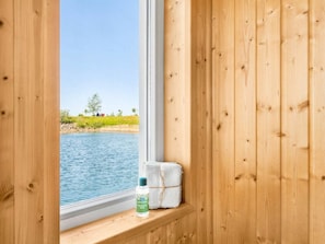A wooden cabin with a window overlooking a body of water and a grassy area. - Holiday Home in Olpenitz With Swimming Pool (Kappeln)