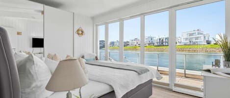 A modern bedroom with a large window, a bed, a bedside table with a lamp, and a view of the water and buildings.
