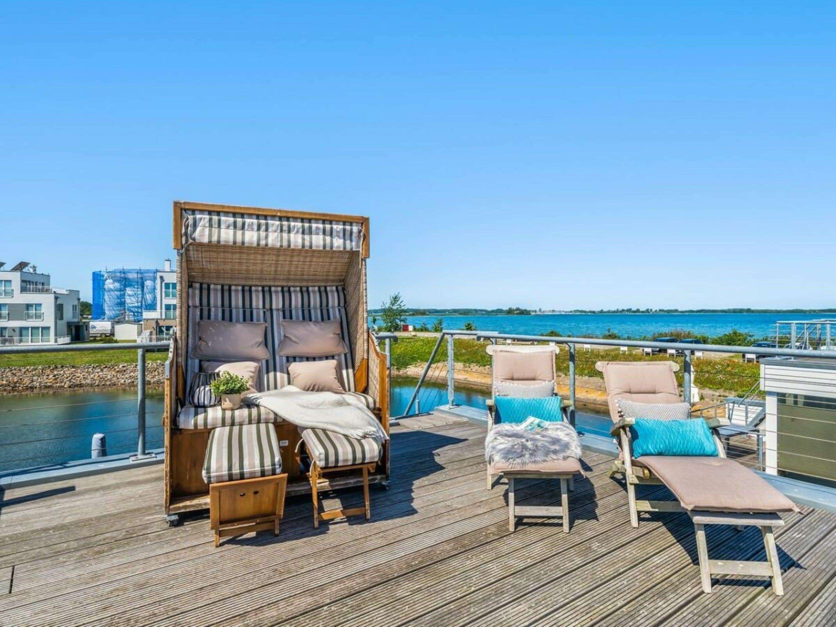 A deck with a wicker canopy and cushioned seating, overlooking a body of water and buildings.