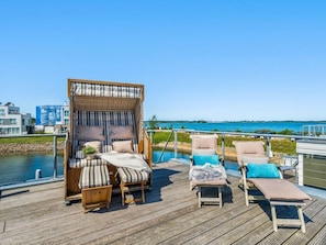 A deck with a wicker canopy and cushioned seating, overlooking a body of water and buildings. - Holiday Home in Olpenitz With Swimming Pool (Kappeln)