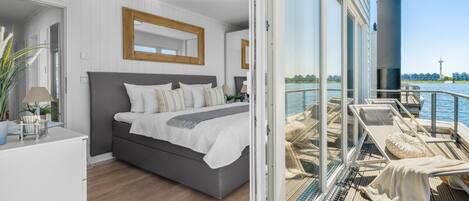 A modern bedroom with a large bed, a view of the water, and a balcony with lounge chairs.