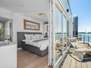Une chambre moderne avec un grand lit, une vue sur l’eau et un balcon équipé de chaises longues.