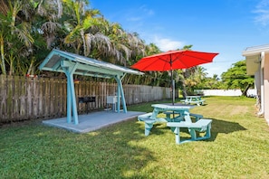 Un jardin avec un patio couvert, des tables de pique-nique et un parasol rouge.