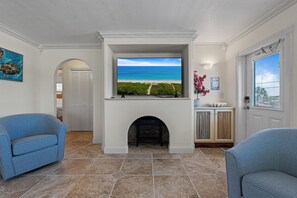A living room with a fireplace, a television displaying a beach view, and two blue armchairs. - Intercoastal Cove Retreat - Upstairs Island Escape with Water Views (Fort Pierce)