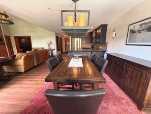 A dining area with a wooden table and chairs, a chandelier, and a kitchen in the background. - Honeycomb Reserve by Cottonwood Lodging (UNDER RENOVATION) (Solitude)
