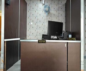 Een receptiebalie met een bordje met daarop 'RECEPTION', een computermonitor en een ventilator aan de muur.