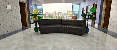 Una habitación amplia con un sofá curvo de color oscuro, dos plantas en macetas, un ventilador de techo y un gran ventanal con vistas a la ciudad.