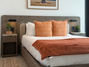 Quarto com cama, almofadas, mesa de cabeceira, luminária na parede e quadro emoldurado acima da cama.