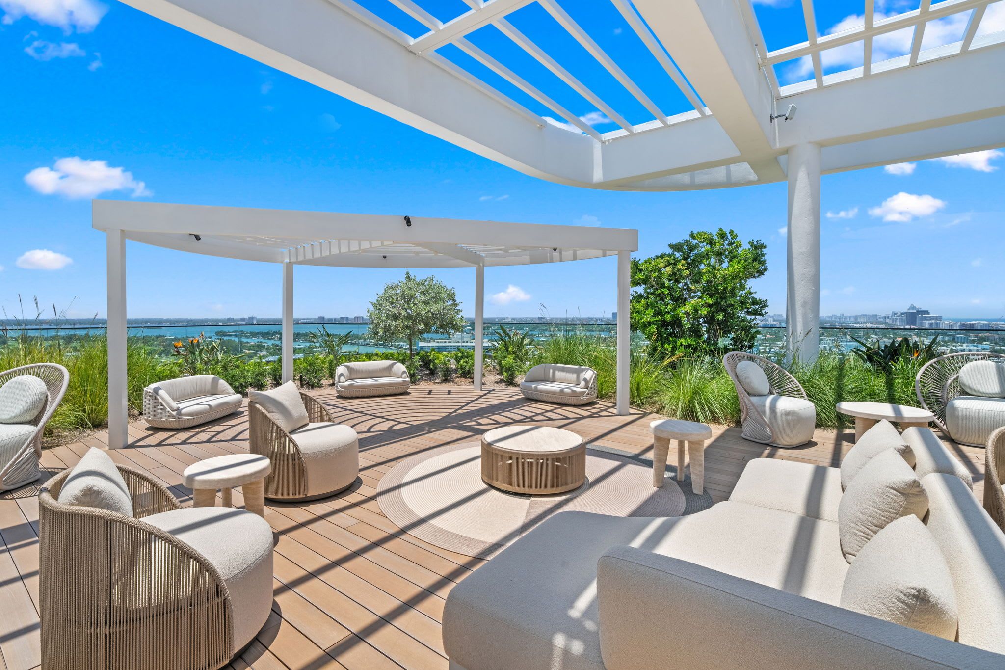 A covered outdoor seating area with multiple lounge chairs and a table, overlooking a body of water and city skyline.