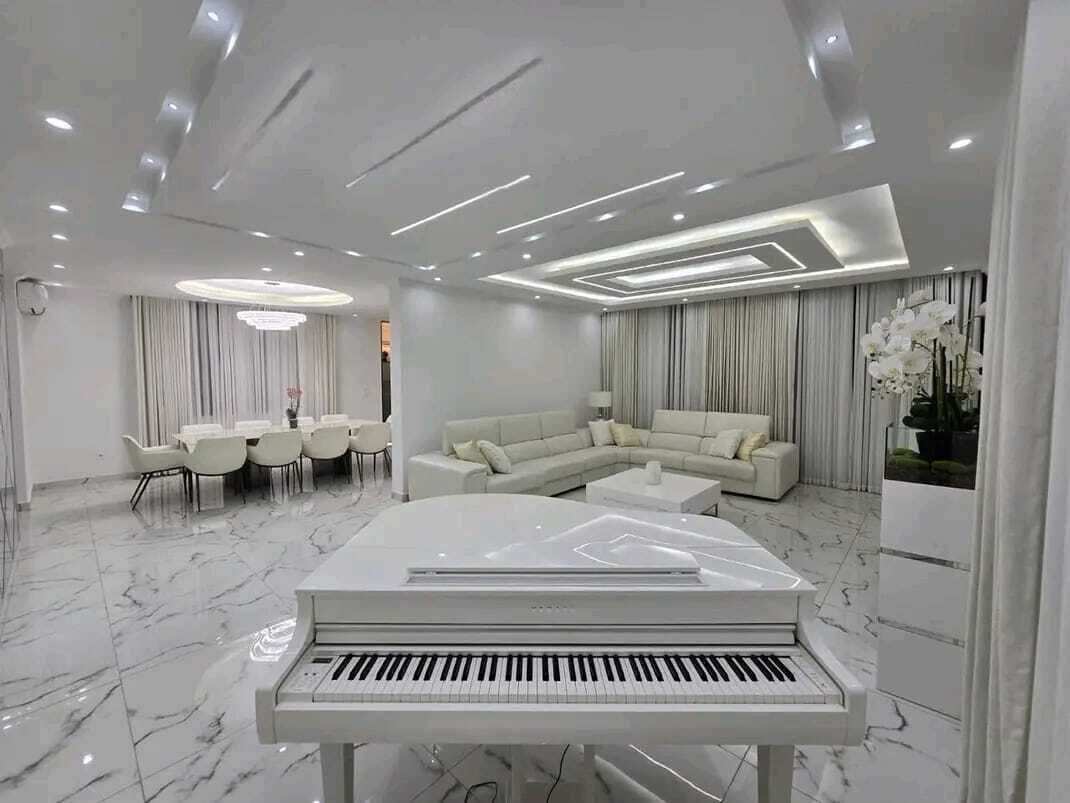 A modern living room with a piano, a sectional sofa, a dining area, and a large window with curtains.