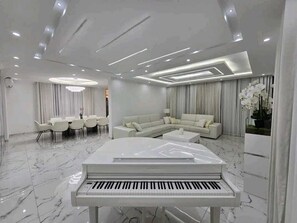 A modern living room with a piano, a sectional sofa, a dining area, and a large window with curtains.
