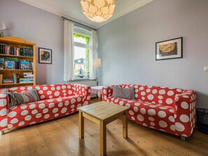A living room with two red sofas with white polka dots, a wooden coffee table, a bookshelf, a window with curtains, and a lamp. - Holiday Apartments Proll (Heringsdorf)