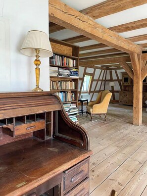 Ein Zimmer mit freiliegenden Holzbalken, einem Holztisch, einer Lampe, einem Bücherregal und einem Stuhl.
