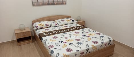 A bedroom with a wooden bed, floral bedding, a nightstand, a bedside table, and a wall-mounted artwork.