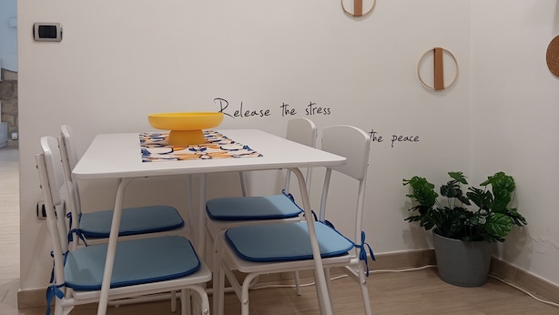 Comedor con una mesa blanca, sillas azules y un tazón amarillo. Se observan adornos en la pared y una planta en maceta.