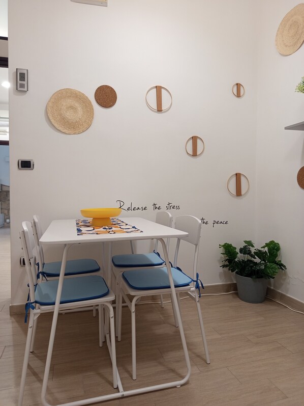 A dining area with a white table, blue chairs, and a yellow bowl. There are wall decorations and a potted plant.