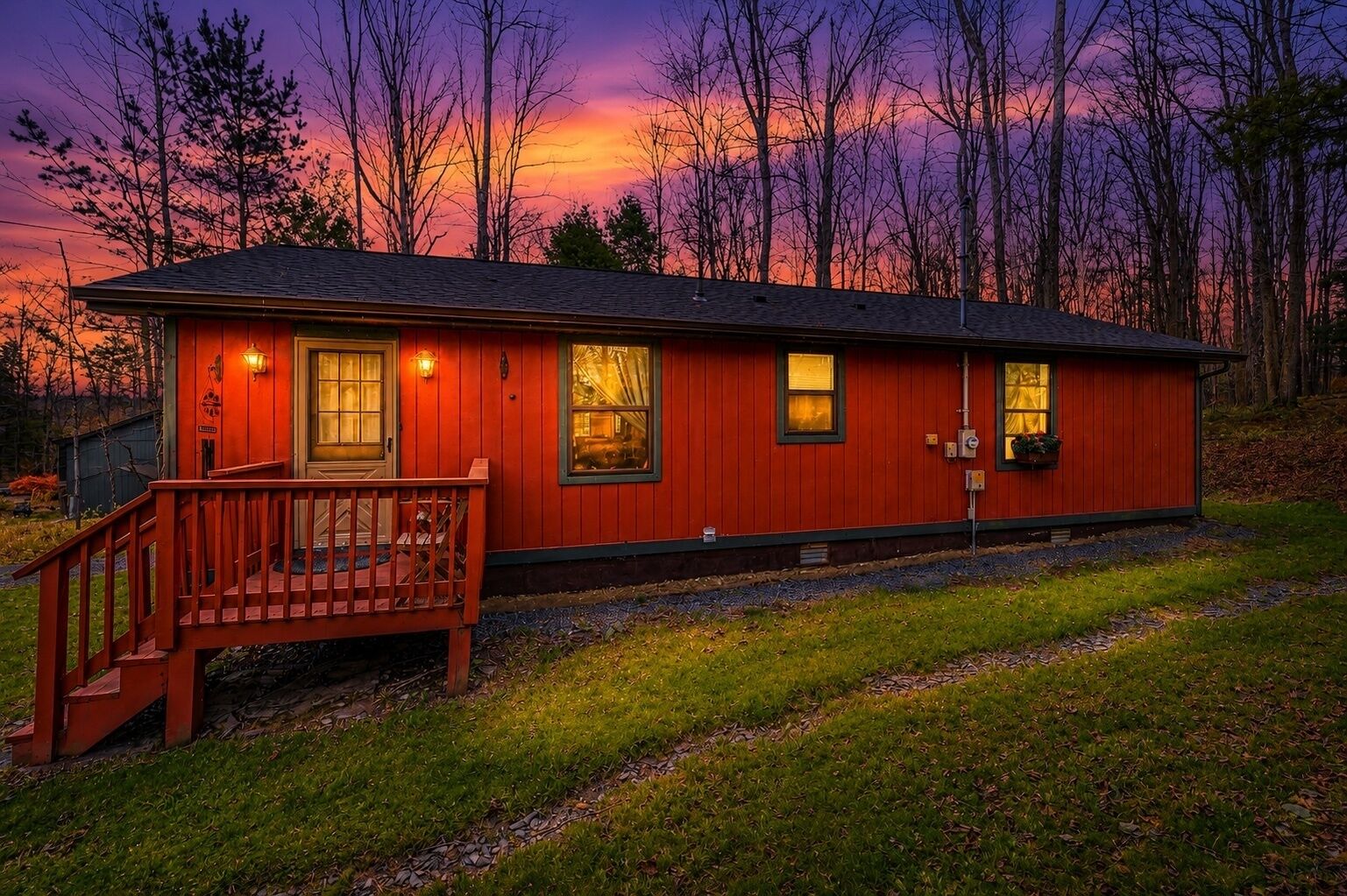 A red cabin with a porch, windows, and a door, surrounded by trees and a sunset sky.