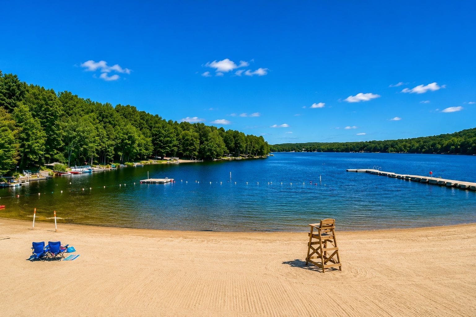 A sandy beach with a lifeguard chair, a clear blue sky, and a calm lake surrounded by trees.