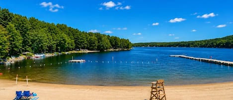 A sandy beach with a lifeguard chair, a clear blue sky, and a calm lake surrounded by trees.