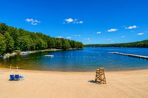 Uma praia arenosa com uma cadeira de salva-vidas, céu azul límpido e um lago calmo rodeado de árvores.