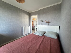 Een slaapkamer met een bed, een radiator, een lamp en een vaas met gedroogde bloemen aan de muur.