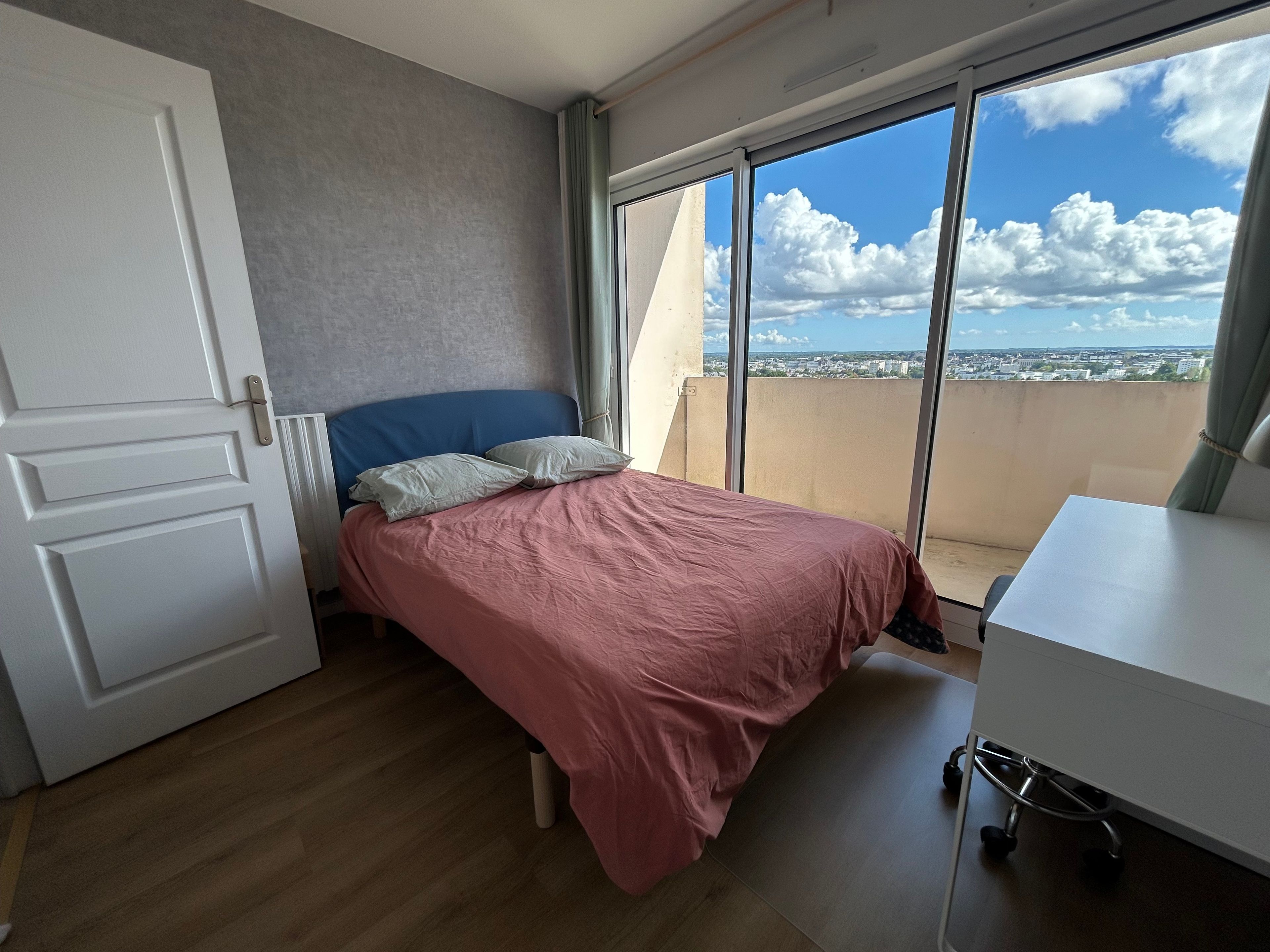 A bedroom with a bed, a desk, a window with a view of the city, and a door.