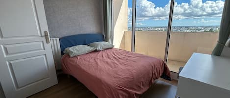 Un dormitorio con cama, escritorio, ventana y vista a la ciudad, además de una puerta.