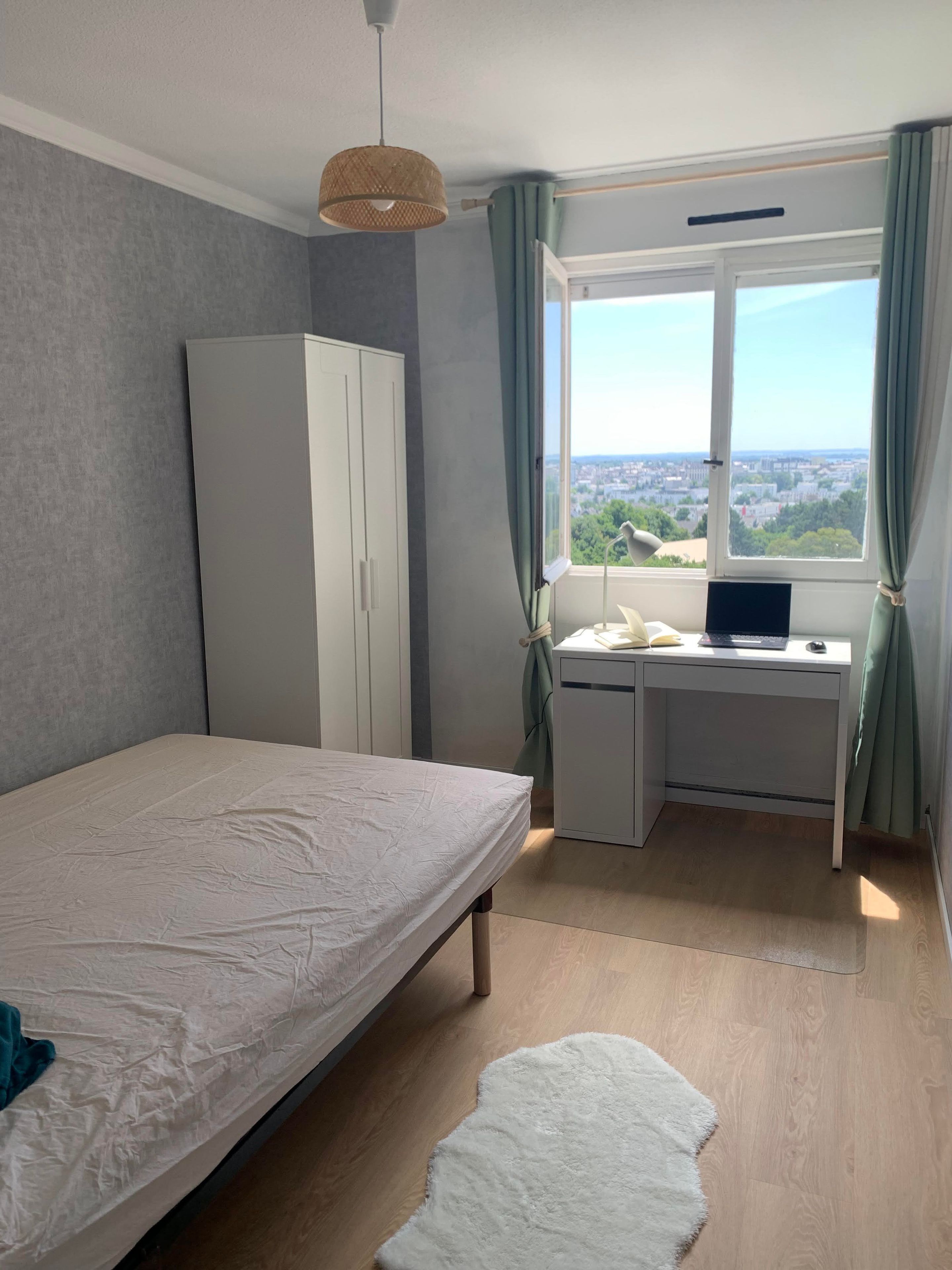 A bedroom with a bed, a desk, and a view of a cityscape through a window.