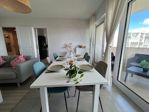 Ein moderner Essbereich mit einem weißen Tisch, gedeckt für vier Personen, umgeben von Stühlen, einer Couch mit gemusterten Kissen und Blick auf einen Balkon mit einer Pflanze.