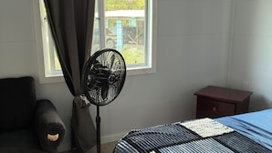 A bedroom with a bed, a standing fan, a chair, a small table, and a window with curtains.