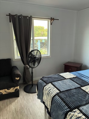 A bedroom with a bed, a standing fan, a chair, a small table, and a window with curtains.