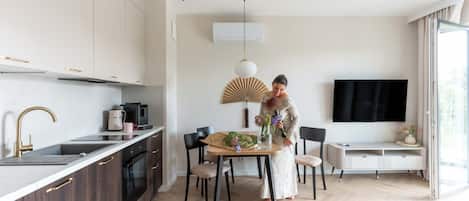 Una cocina moderna con horno empotrado, cafetera y lavaplatos. Hay una zona de comedor con mesa y sillas, y un televisor montado en la pared.