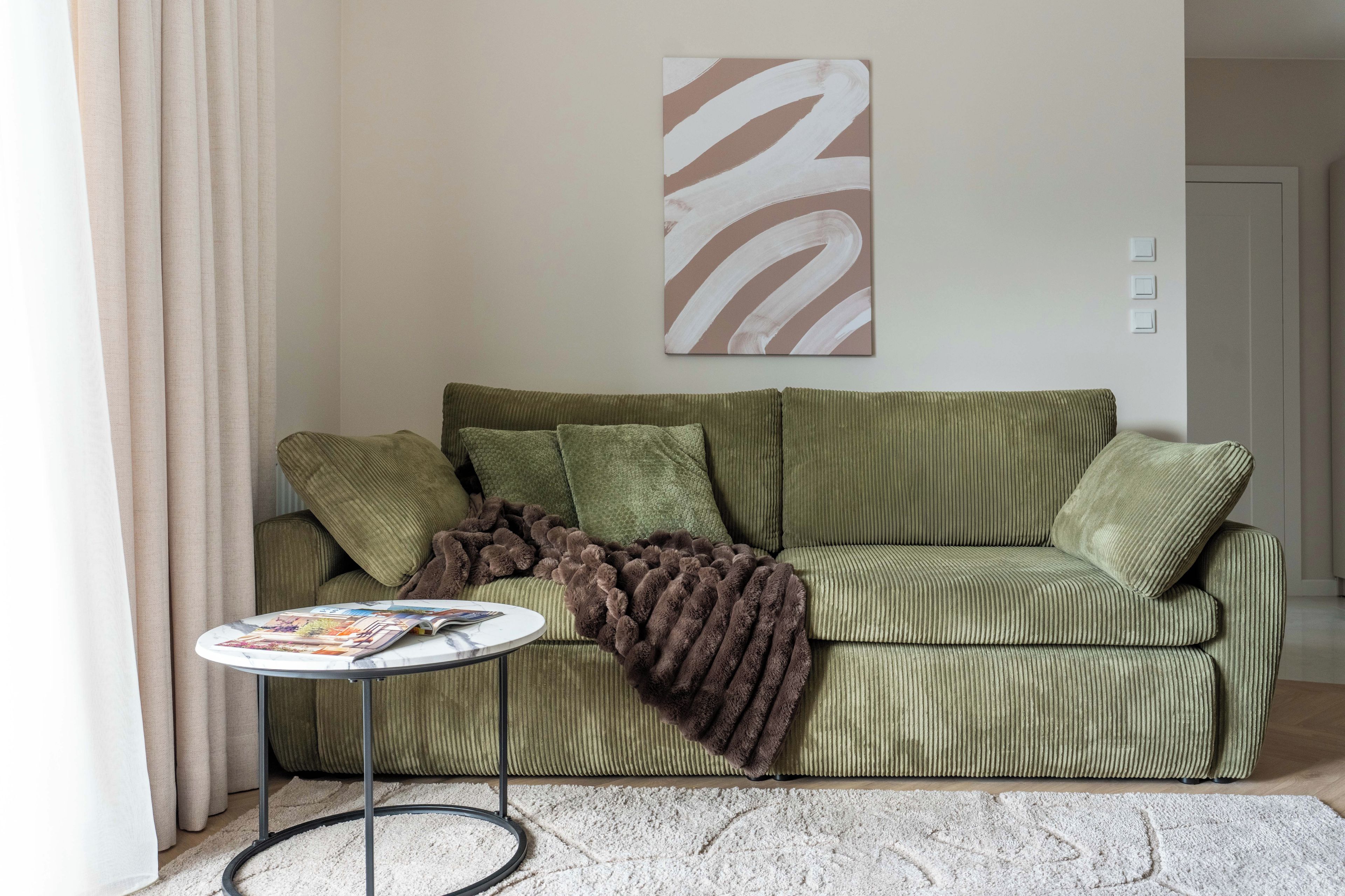 Un canapé vert avec un plaid marron, une petite table ronde sur laquelle sont posés des magazines, et un grand tableau abstrait accroché au mur.