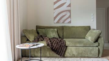 Un canapé vert avec un plaid marron, une petite table ronde sur laquelle sont posés des magazines, et un grand tableau abstrait accroché au mur.
